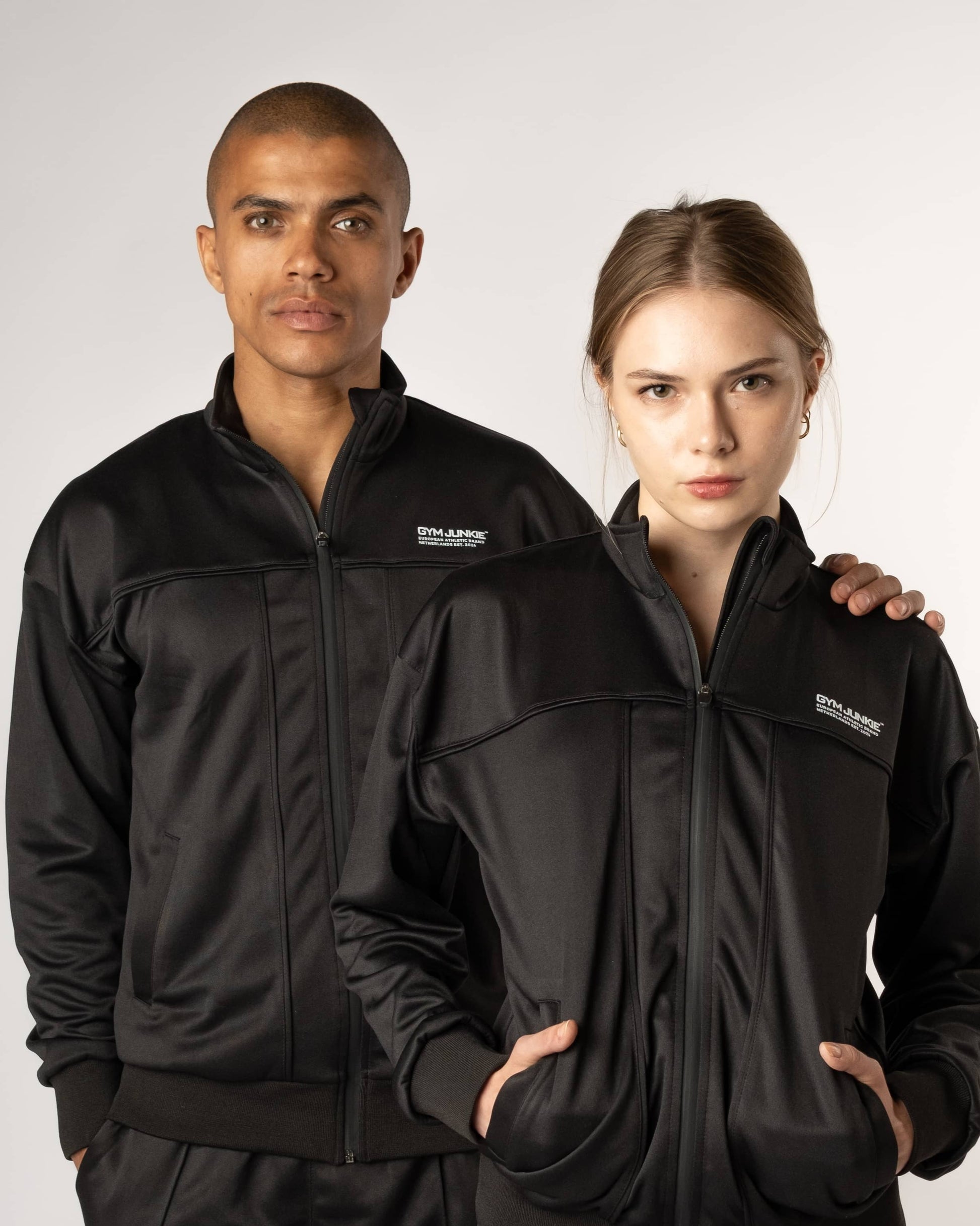 Man en vrouw dragen GJA uniseks trainingsjas, stijlvolle en comfortabele sportjassen voor jonge, gedurfde sporters in Nederland - Gym Junkie Sportkldeing