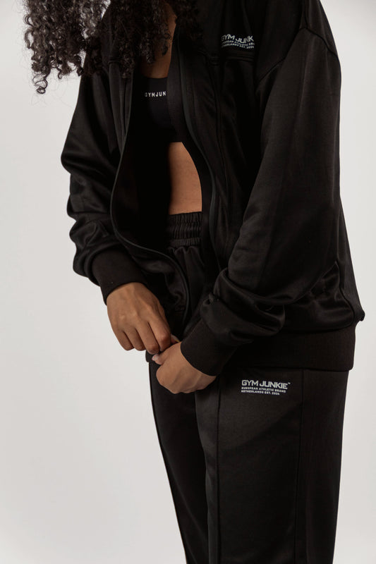 Model ritst GJA trainingsjas dicht, met bijpassende trainingsbroek, stoere en comfortabele gymoutfit voor jonge, zelfverzekerde vrouwen in Nederland 
- Gym Junkie Sportset voor dames